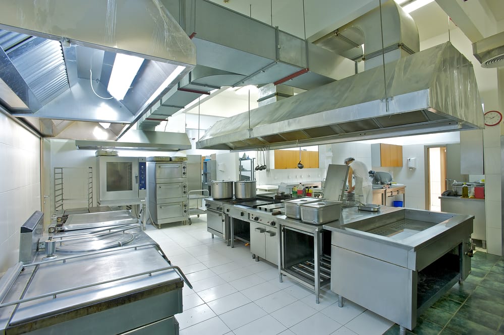 Spacious commercial kitchen with large overhead ventilation ducts and stainless steel equipment, showcasing an effective AC unit in a commercial kitchen for airflow and heat control.