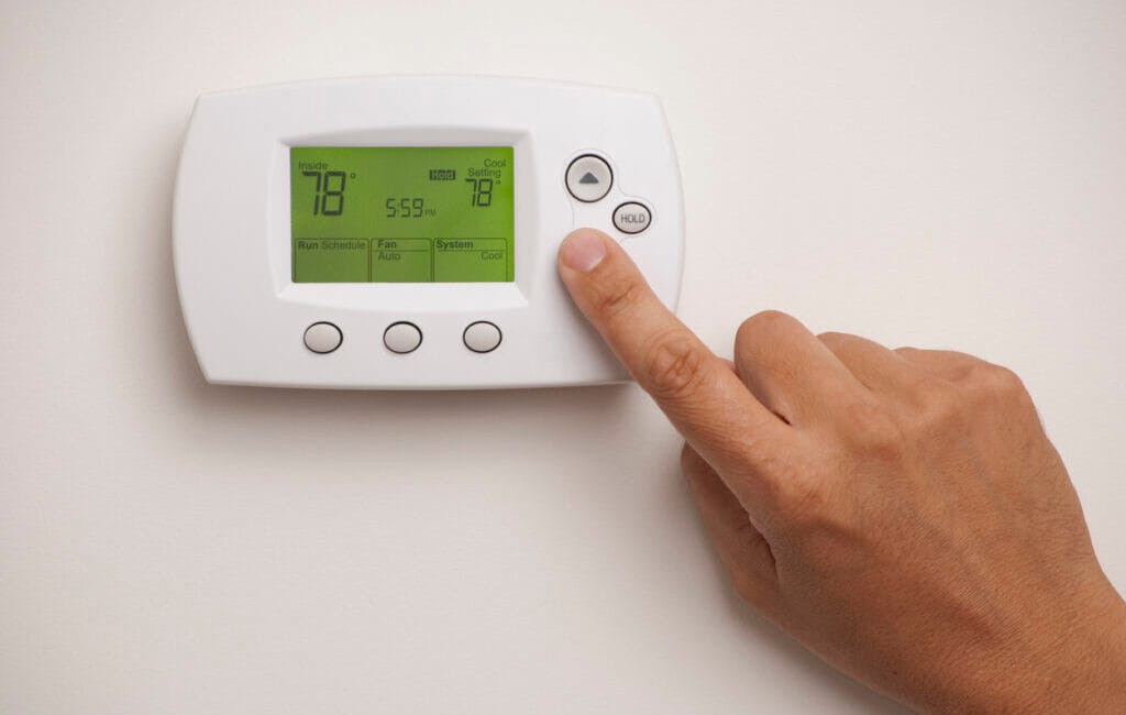 A person pressing the “up” button on a digital thermostat, possibly adjusting settings to turn off HVAC when it isn’t heating.
