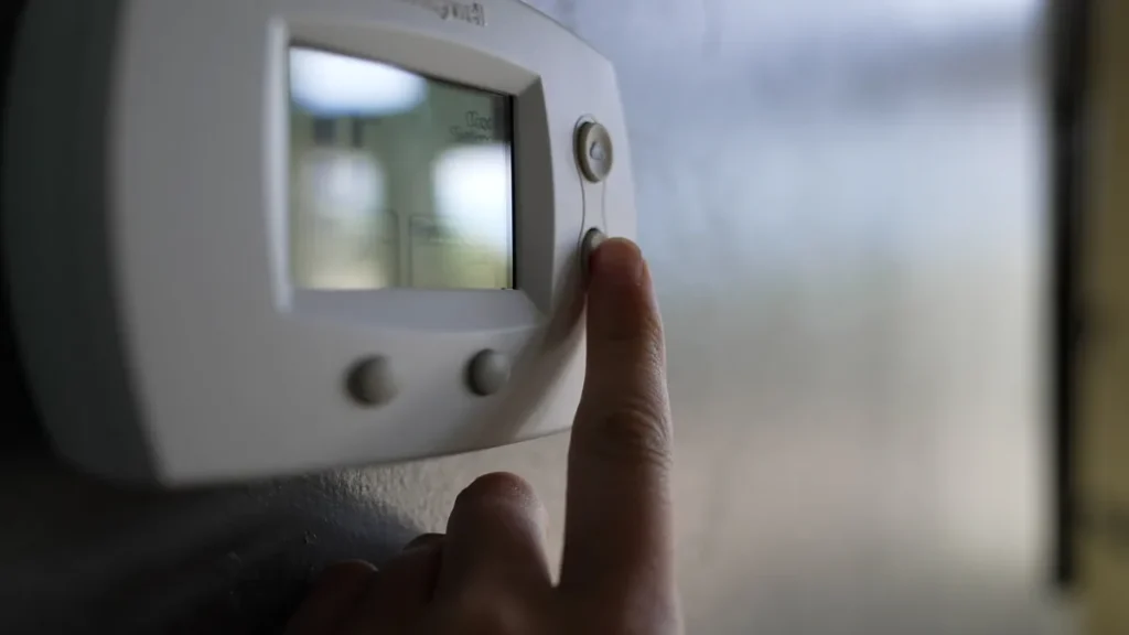 Close-up of a hand changing the temperature on a home thermostat — a step often taken to turn off HVAC when heating isn’t needed.