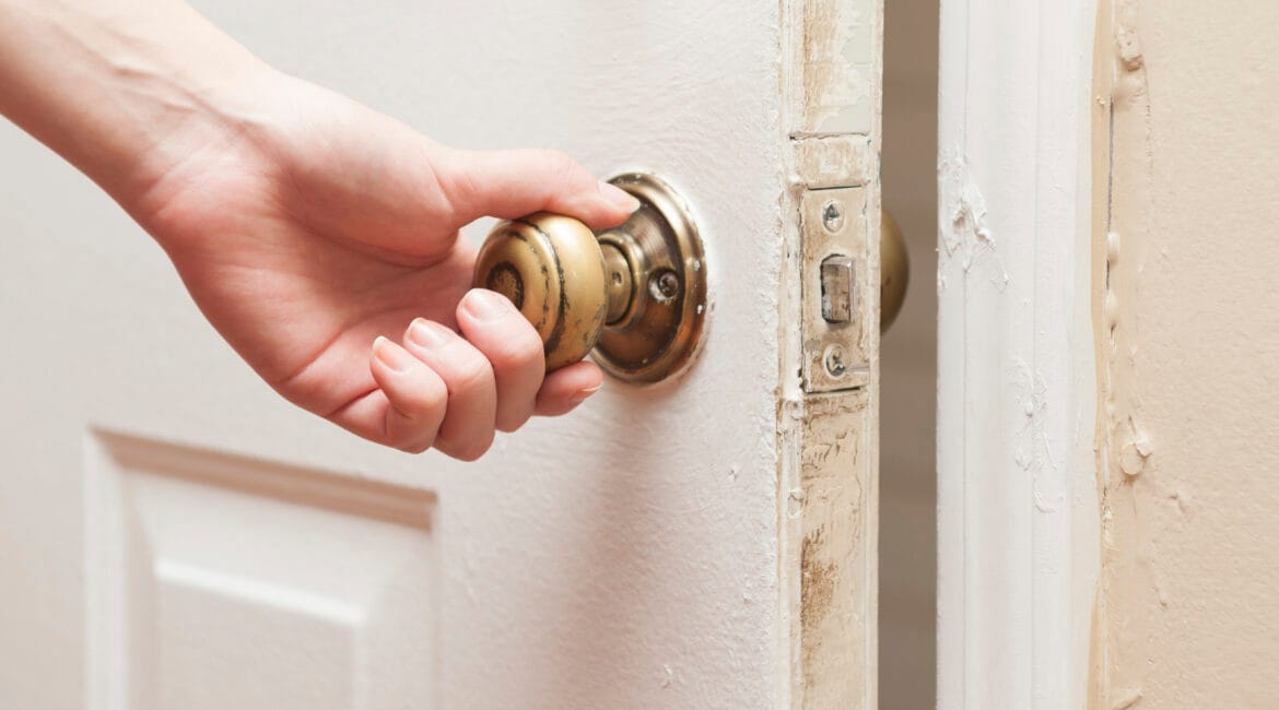 Close-up of a hand turning an old brass doorknob on a partially opened door, representing the decision to leave doors open or closed in home settings.