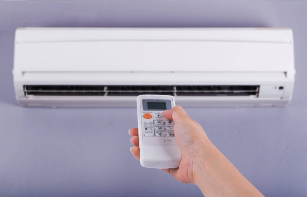 Hand holding remote control to operate wall-mounted air conditioner unit.
