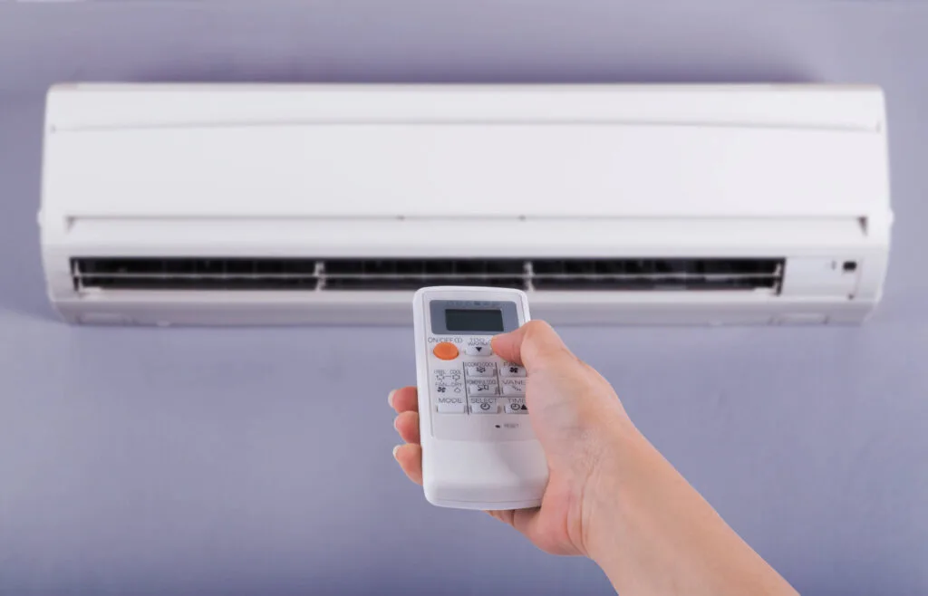 Hand holding remote control to operate wall-mounted air conditioner unit.