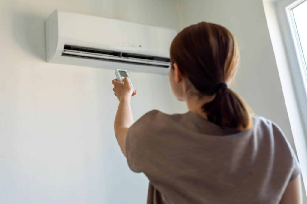 Woman pointing a remote control at wall-mounted air conditioner, turning her AC up when she's not home.
