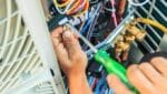 Hands of a technician using a screwdriver to adjust wiring inside an outdoor air conditioning unit during aircon servicing.