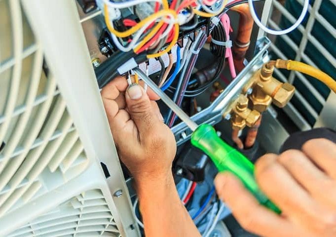 Hands of a technician using a screwdriver to adjust wiring inside an outdoor air conditioning unit during aircon servicing.
