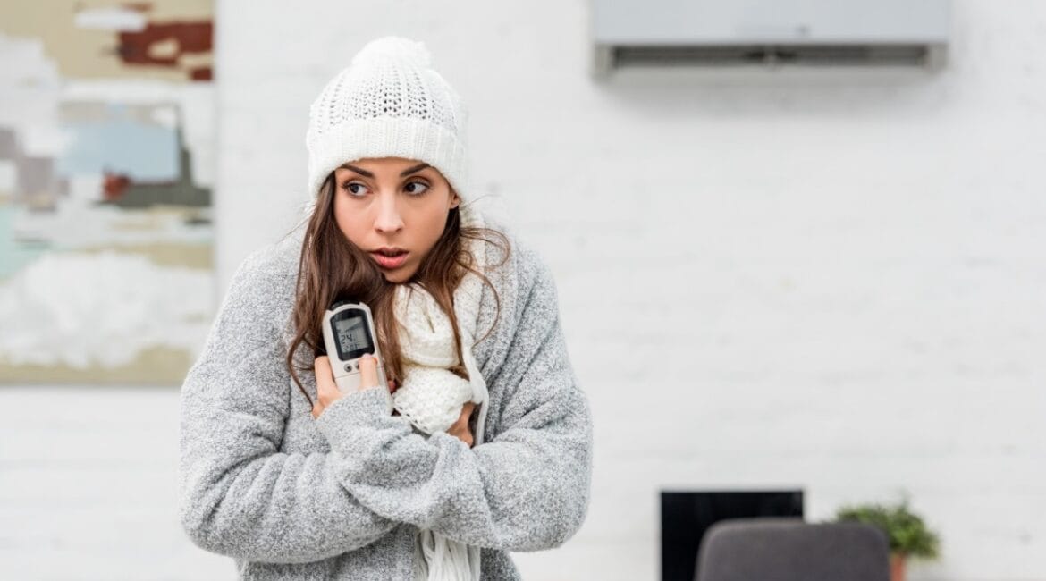 A woman bundled in winter clothes struggles to heat her home, highlighting the need for a more efficient heating solution.