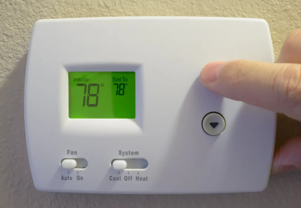Finger pressing a button on a wall thermostat, which can lead to heating mistakes if not programmed correctly.