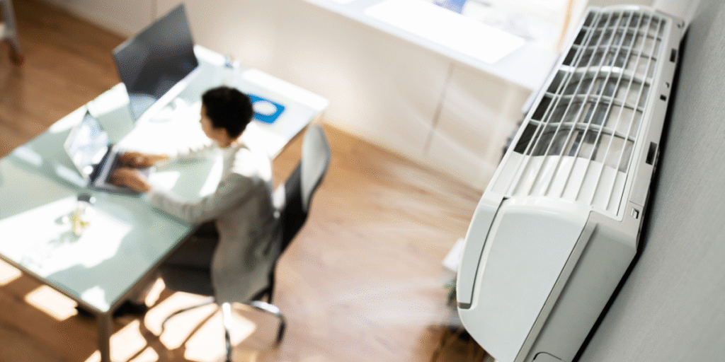 Office worker sitting under a running air conditioner during a heatwave — demonstrating how to avoid thermal shock when using air conditioning in severe weather through proper temperature settings and professional air conditioning Gold Coast maintenance.