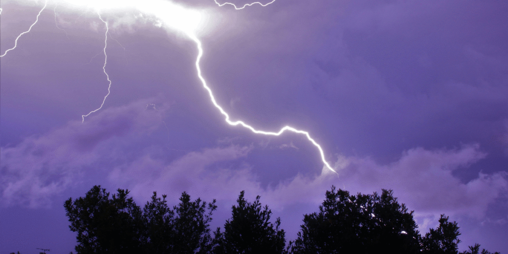 Bright lightning bolt hitting near residential trees under stormy clouds, illustrating how severe weather can cause an air conditioner struck by lightning to stop working. Useful visual for air conditioning repair service Gold Coast storm safety awareness.