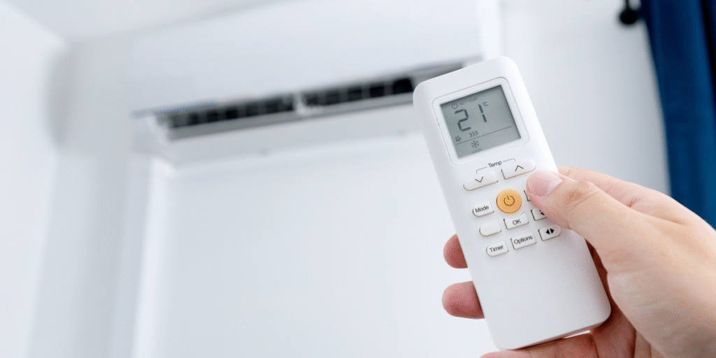 Hand holding a remote control to turn on an overloaded air conditioner in hot weather, illustrating proper use of air conditioning Gold Coast systems.