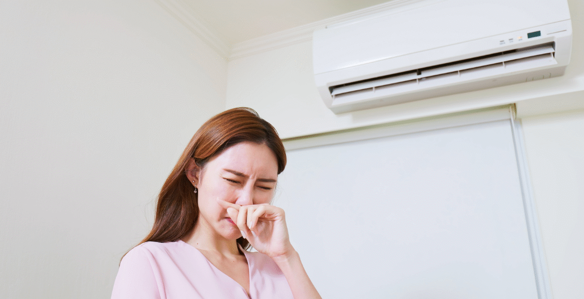 Woman reacting to strong air conditioner smell coming from a wall-mounted unit, showing common indoor comfort issues with air conditioning Gold Coast systems.