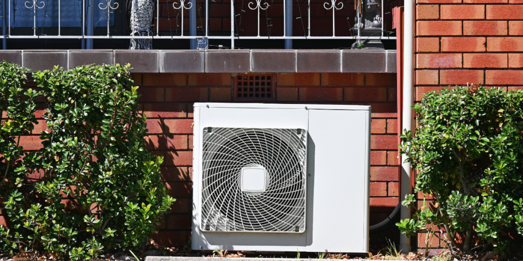 Rusty outdoor air conditioner unit placed beside a house wall with green bushes around, illustrating corrosion issues common in coastal climates — air conditioning Gold Coast.