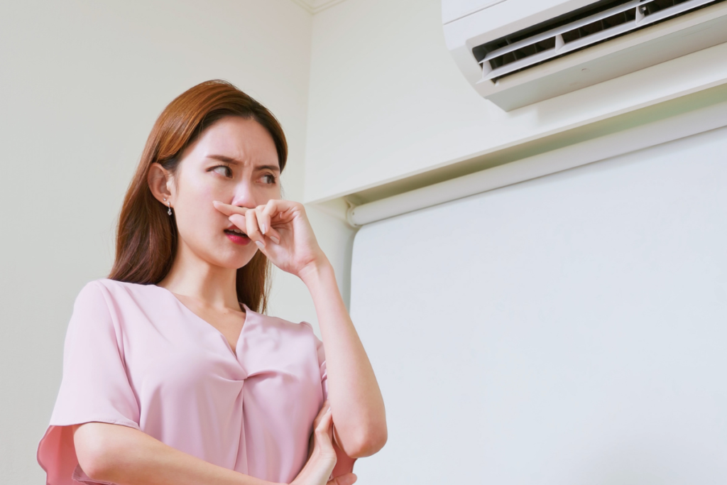 Woman reacting to unpleasant smell from wall-mounted unit, illustrating autumn comfort problems and what is the common problem with an air conditioner in fall, air conditioning Gold Coast.