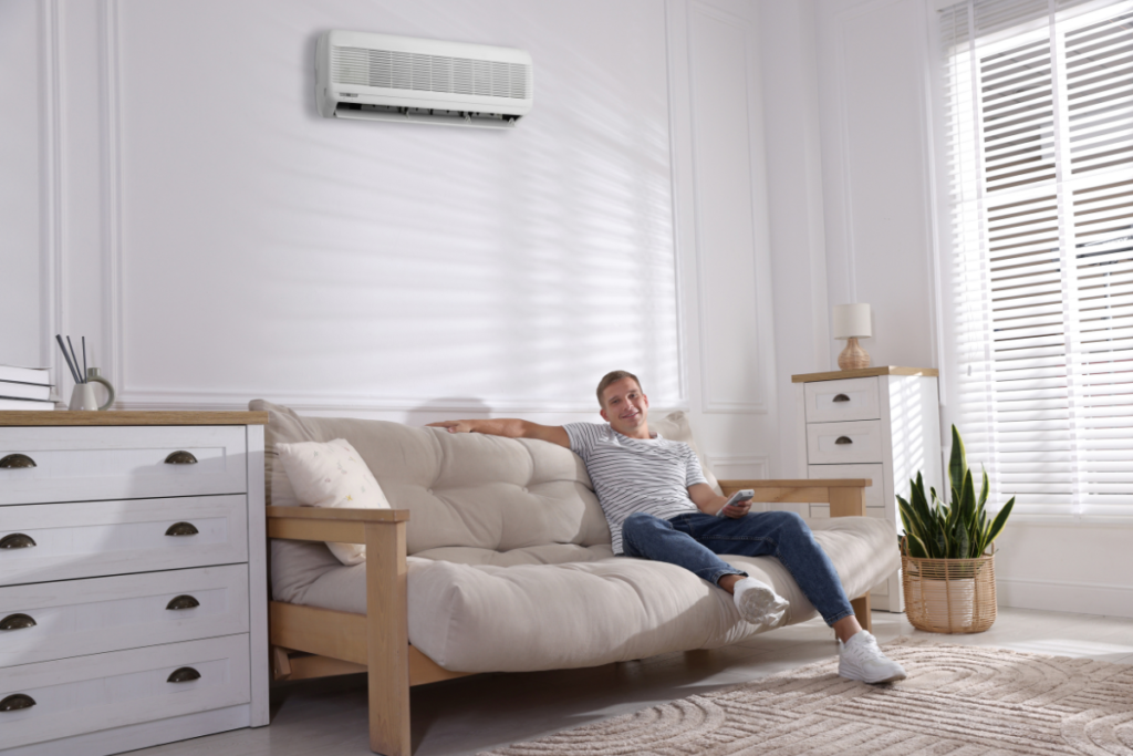 Homeowner relaxing under a wall-mounted air conditioner during summer, highlighting what to consider before replacing an AC in heatwave conditions.