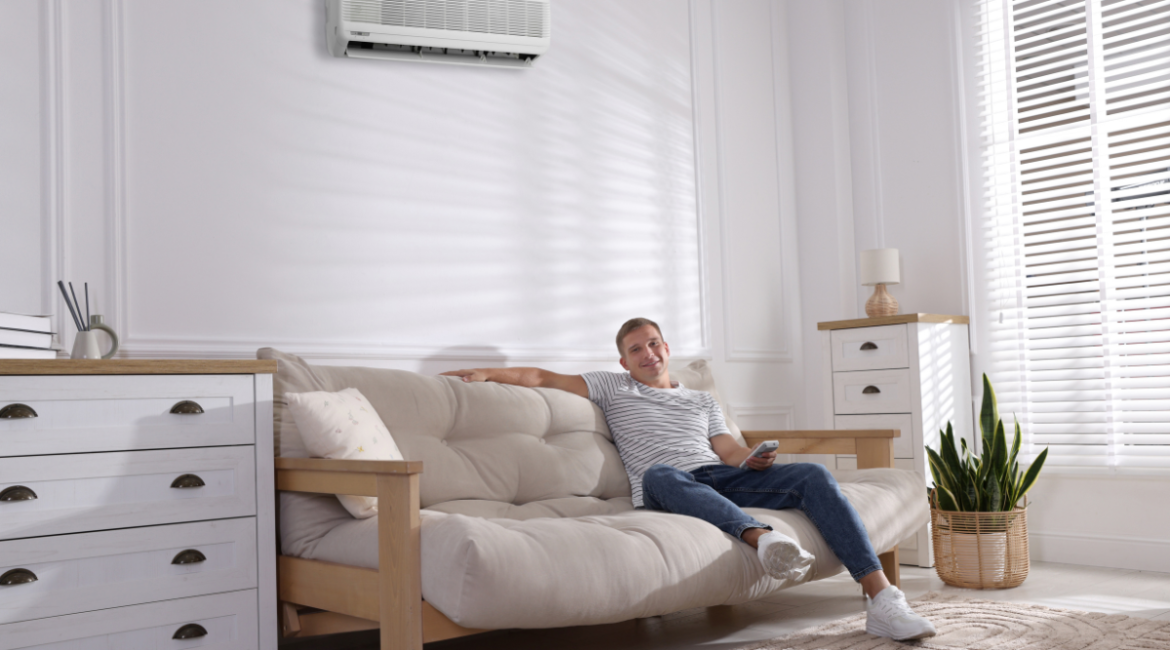 Homeowner relaxing under a wall-mounted air conditioner during summer, highlighting what to consider before replacing an AC in heatwave conditions.