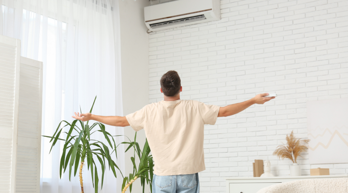 Man standing under wall-mounted air conditioner using remote, demonstrating AC dry mode for better humidity control in residential air conditioning Gold Coast homes, showing if it is cheaper to run AC on dry mode