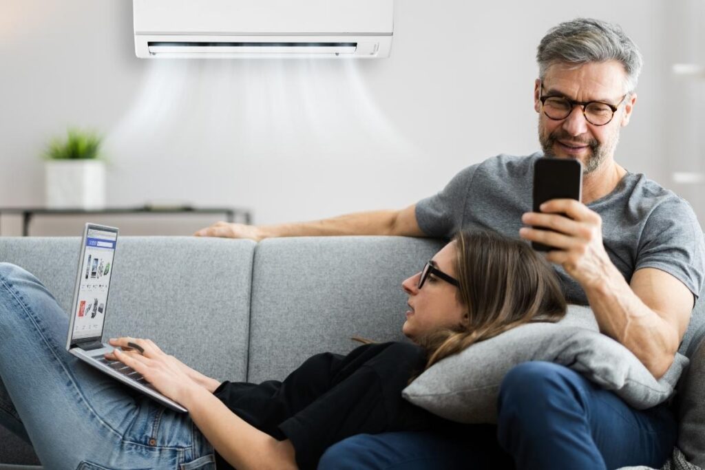 A couple relaxing on a sofa under a split system unit, wondering does air conditioning make the air too dry in their air conditioning Gold Coast home.
