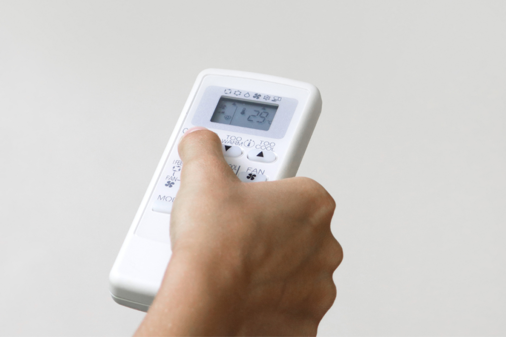 Hand holding and pressing buttons on a white AC remote, specifically highlighting 'TOO WARM' and 'TOO COOL' options. Adjusting settings brings up the question, 'Do air conditioners make the room more dry?' while using a system, a key consideration for healthy use that can make air dry.