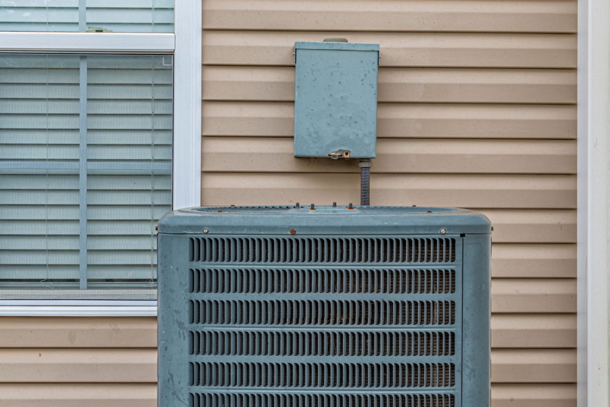 A residential air conditioning condenser unit installed outside a house with beige siding, highlighting the importance of regular air conditioning maintenance gold coast to keep electricity bills low.