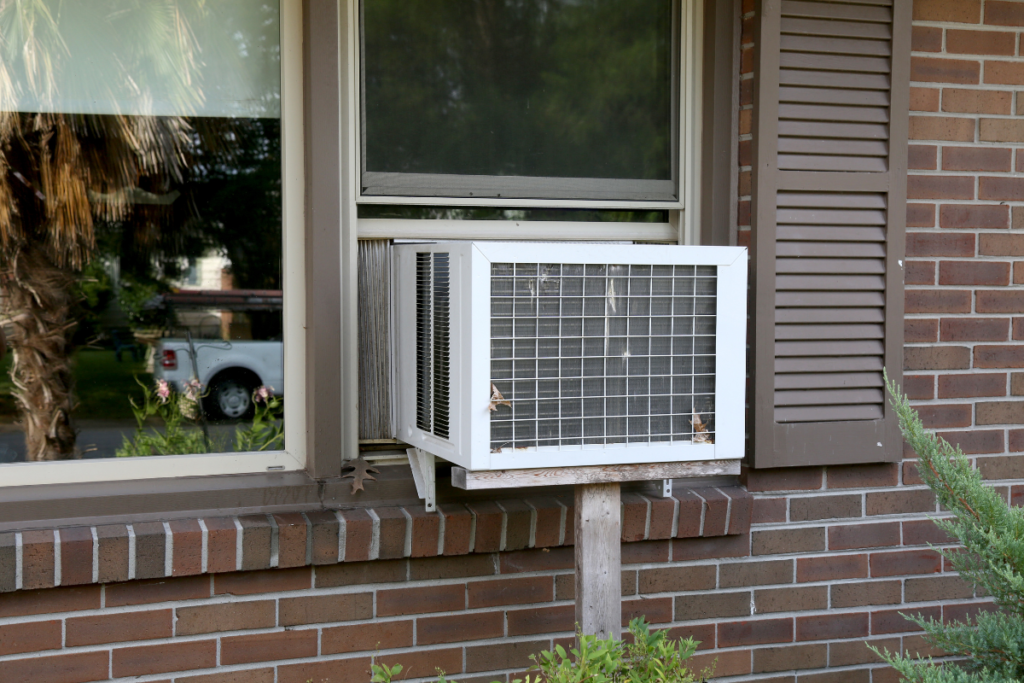 Close-up of an old-school residential air conditioning gold coast window unit supported by a wooden post, demonstrating why routine upkeep is essential to lower your aircon electricity bill.