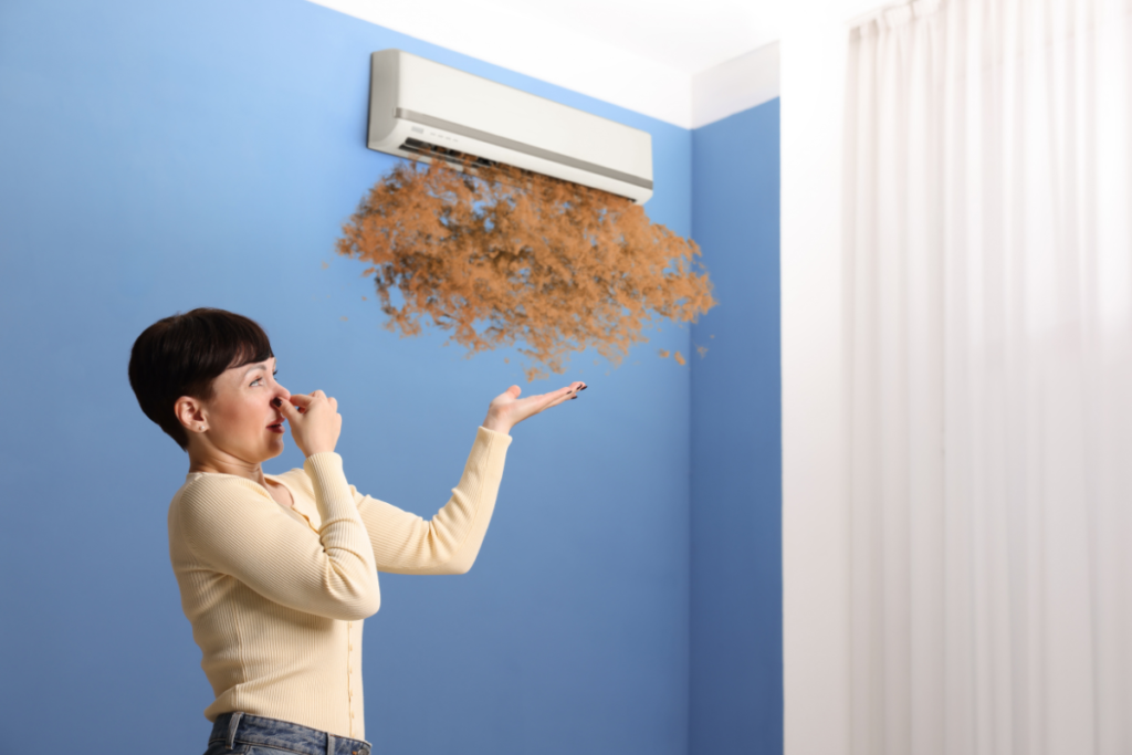 A woman holds her nose and winces as brown, dust-like particles come out of a wall-mounted air conditioning unit, showing a serious problem where the aircon smells bad.