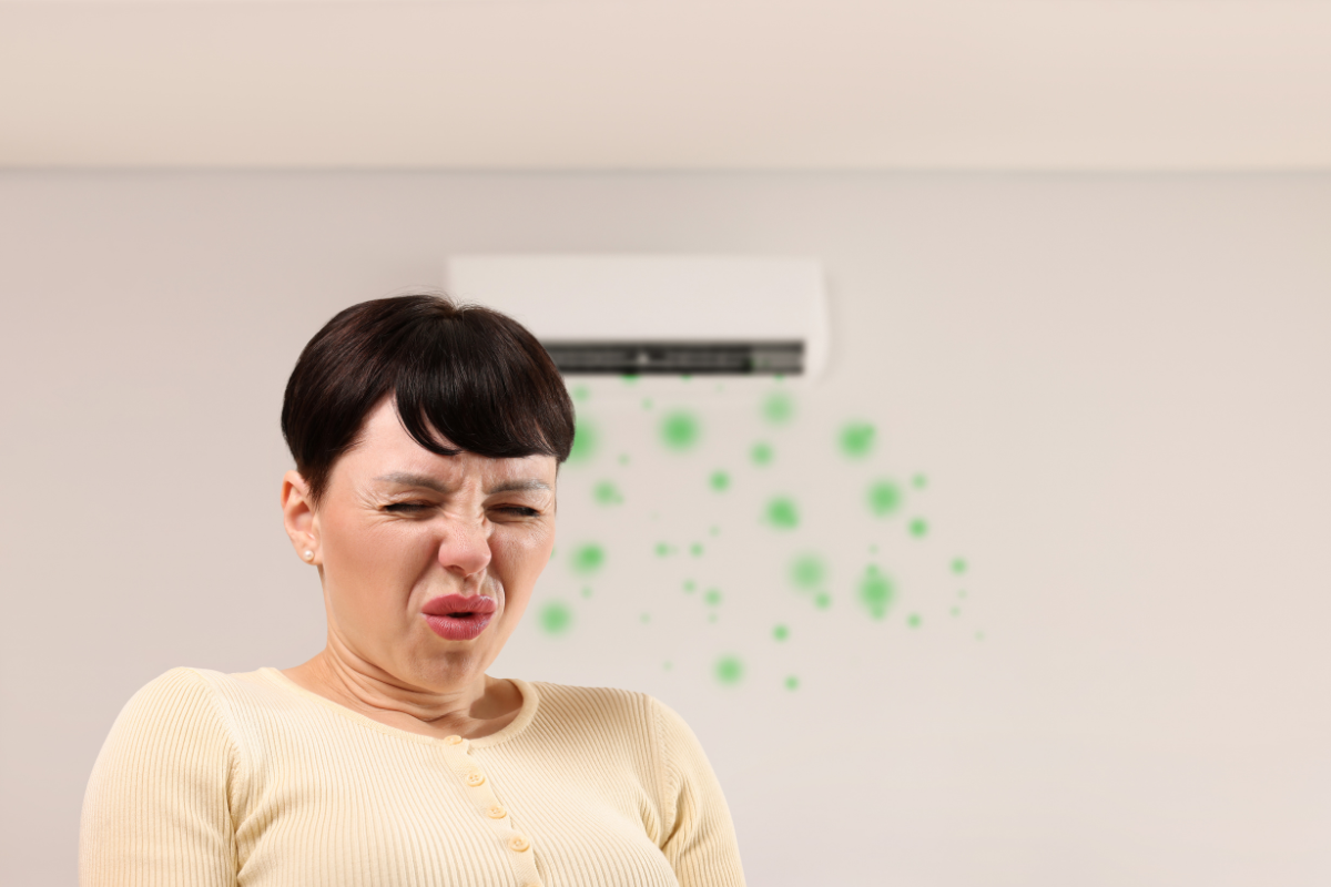 A woman makes a disgusted face, holding her nose as a wall-mounted split system air conditioner blows a plume of green, dusty air toward her, often happening when the aircon smells bad when heating.