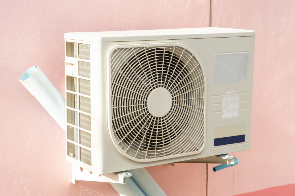 A white residential air conditioning gold coast outdoor unit mounted on brackets against a pastel pink wall, properly serviced to ensure it runs efficiently without producing any unusual white steam.