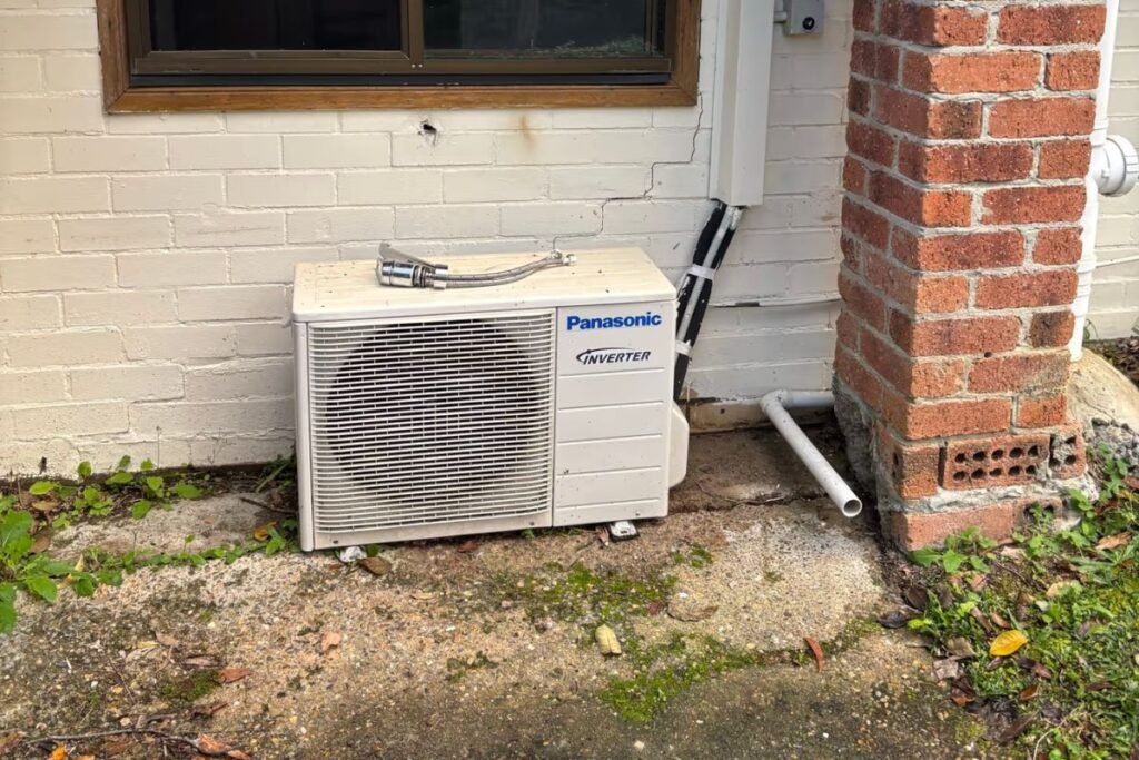 Ground-level outdoor unit surrounded by weeds and leaves that need clearing before an aircon service